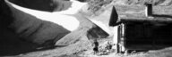 Schwarz-Weiss-Fotografie. In einer Berglandschaft im Vordergrund eine Berghütte auf einer Wiese mit zwei Menschen. Im Hintergrund zwischen Bergen ein Gletscher. - vergrösserte Ansicht