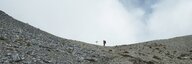 Blick auf eine steinige Landschaft, am Horizont steht nur noch klein sichtbar eine Person neben einem Wegweiser. Darüber wolkiger Himmel. - vergrösserte Ansicht