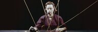 Zum Titel «Die Kunst des Netzwerks» ist eine kunstvoll inszenierte Fotografie aus den 1950ern gewählt. Das Bild zeigt eine Frau mit Headset vor schwarzem Hintergrund. Die Frau hat nach hinten gebundene, braune Haare. Sie trägt eine dunkelrote Bluse und eine enge Halskette. Sie sitzt frontal an einem schwarzen Holztisch, direkt vor ihr eine flache Konsole mit Knöpfen und Kabeln. Die Kabel kommen von ausserhalb des Bildes, verlaufen diagonal und überschneidend und bündeln sich in der Konsole - vergrösserte Ansicht