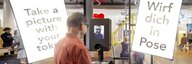Ein Mann steht vor einem Selfieautomaten. Um ihn herum sind Leuchtschirme aufgebaut auf denen anspornende Sätze stehen wie: Wirf dich in Pose. - vergrösserte Ansicht