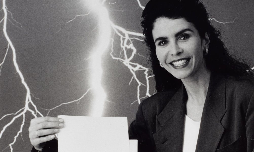 A black and white photo of a laughing woman pulling a sheet of paper out of an old fax machine. A flash of lightning strikes in the background.