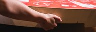 A hand grips a large red table. On it a labyrinth and drawings. In the background, another person can be seen moving the table. - enlarged view