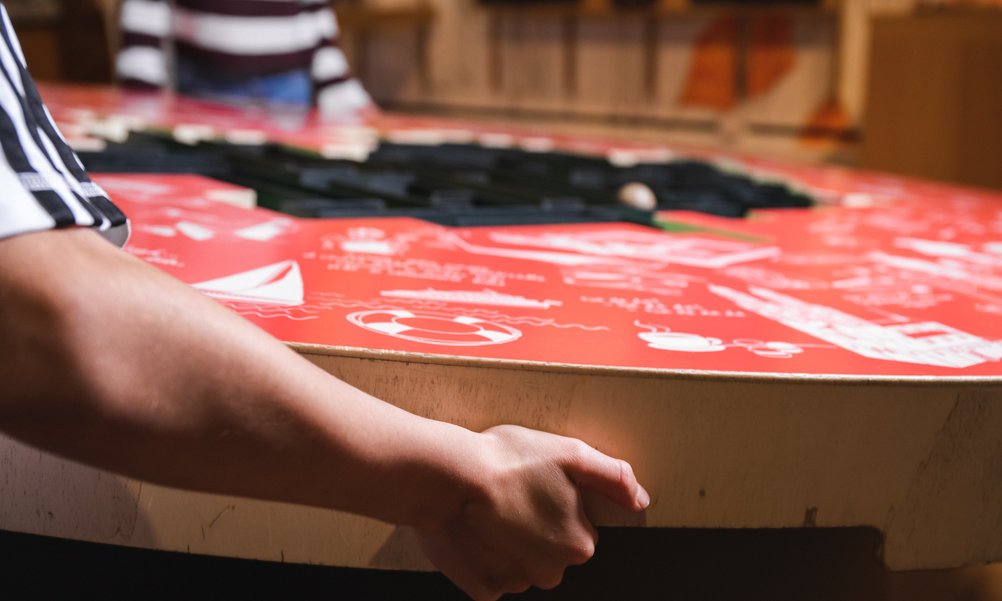 A hand grips a large red table. On it a labyrinth and drawings. In the background, another person can be seen moving the table.