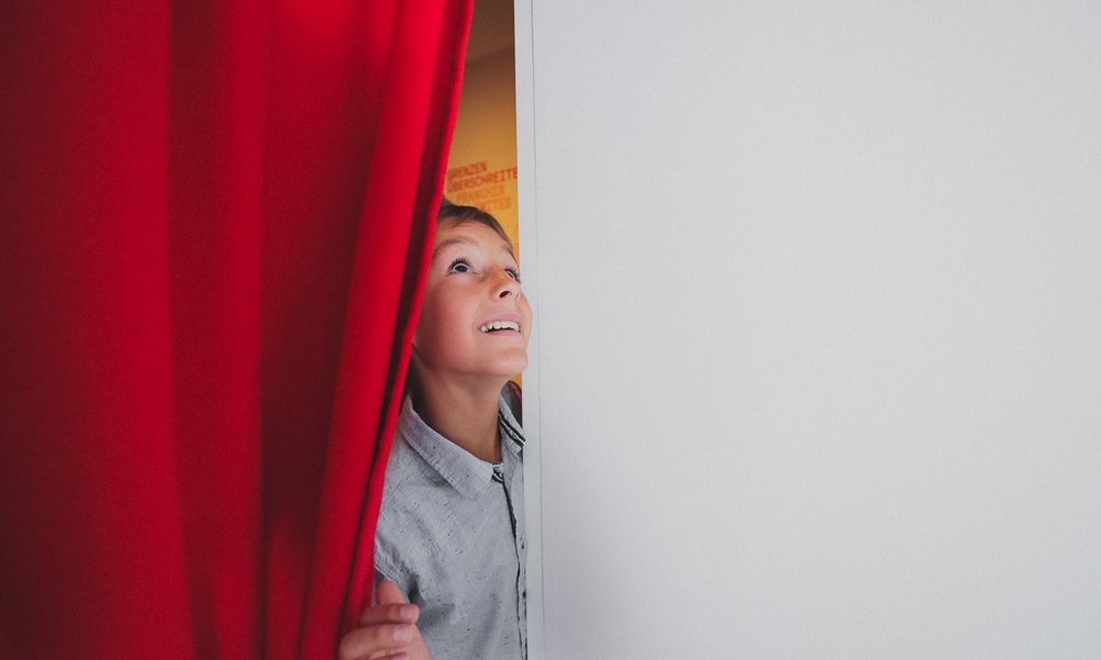Ein Kind schaut hinter einem roten Vorhang hervor fasziniert nach oben.