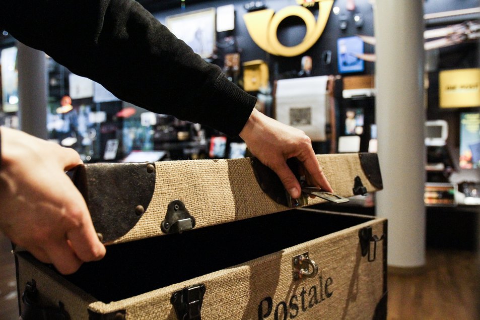 Postkiste Eine Hand öffnet in der Ausstellung eine grosse Post-Kiste. Im Hintergrund sind Ausstellungsobjekte zu sehen.