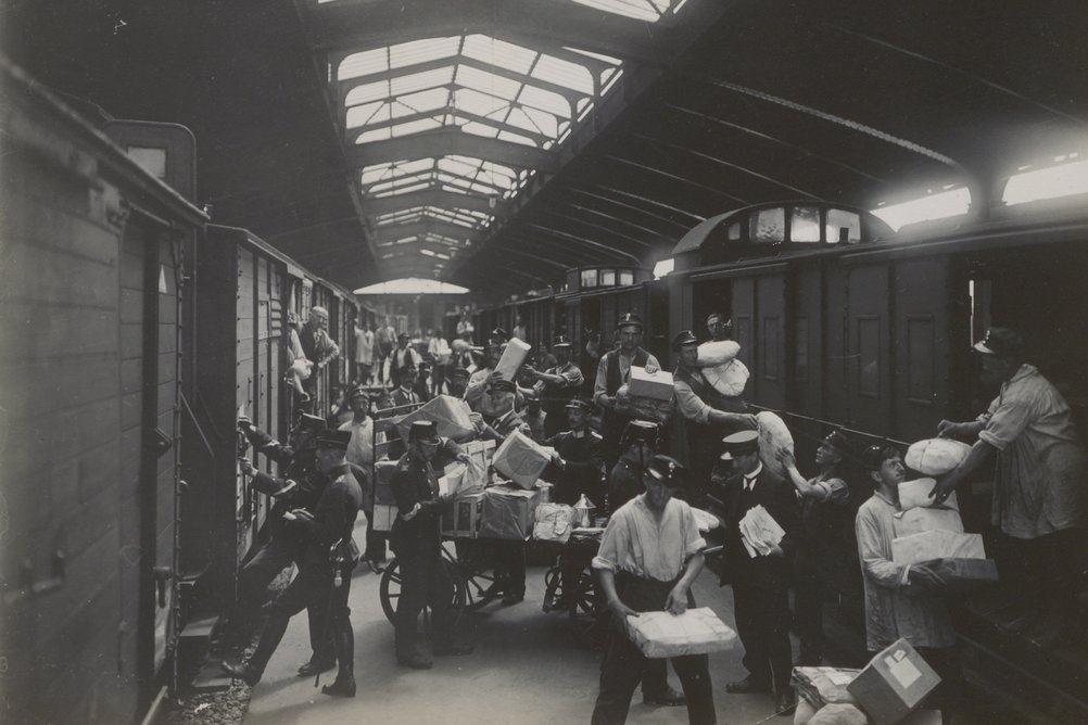 Ein historisches schwarz-weiss Foto von zwei Postzügen, dazwischen wimmelt es von Postmitarbeitenden, die eifrig Pakete umladen.