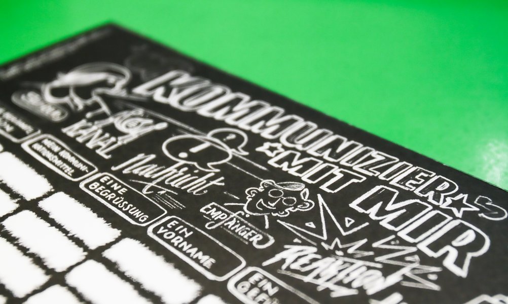 Auf grünem Hintergrund liegt ein schwarz-weisses Spielblatt mit Zeichnungen und Feldern zum Ausfüllen.