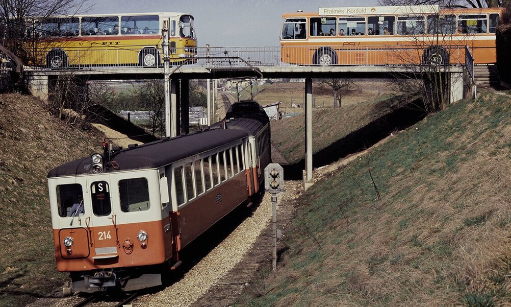 Ein historisches Foto aus dem Jahr 1979: Im Vordergrund ein Zug, etwas zurückversetzt auf einer Brücke über dem Zug kreuzen sich ein Postauto und ein oranger Bus. Im Hintergrund Berge.
