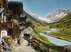 Nachkolorierte Fotografie eines Bergdorfes: Links sind dunkle Holzhäuser zu sehen, davor ein Weg. Etwas weiter rechts fliesst der Fluss Lonza vorbei, im Hintergrund sind Schneeberge zu sehen.