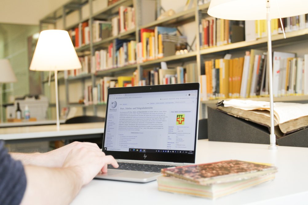 Sur la table de la salle de lecture des archives des PTT se trouve un ordinateur portable avec Wikipedia ouvert. Une personne s'occupe de l'ordinateur portable.