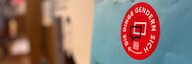 Nahaufnahme eines roten Klebers mit dem Text "Die Dinge gendern sich" auf einem blauen Plakat - dahinter unscharf die Ausstellung. - vergrösserte Ansicht