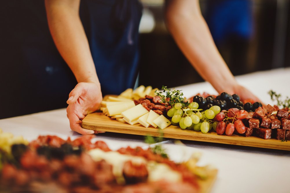 Zwei Hände stellen eine schön angerichtete Holzplatte mit Käse und Früchten aufs Buffet.