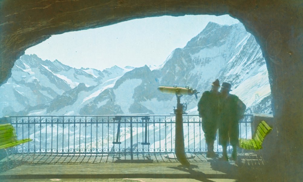 Photographie historique d'une fenêtre de galerie avec télescope. Deux personnes regardent le panorama de la montagne.