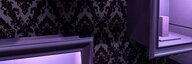 In einem dunklen Raum mit gemusterter Tapete an der Wand sind violette, leere Boxen zu sehen, die von innen ausgeleuchtet sind. - vergrösserte Ansicht