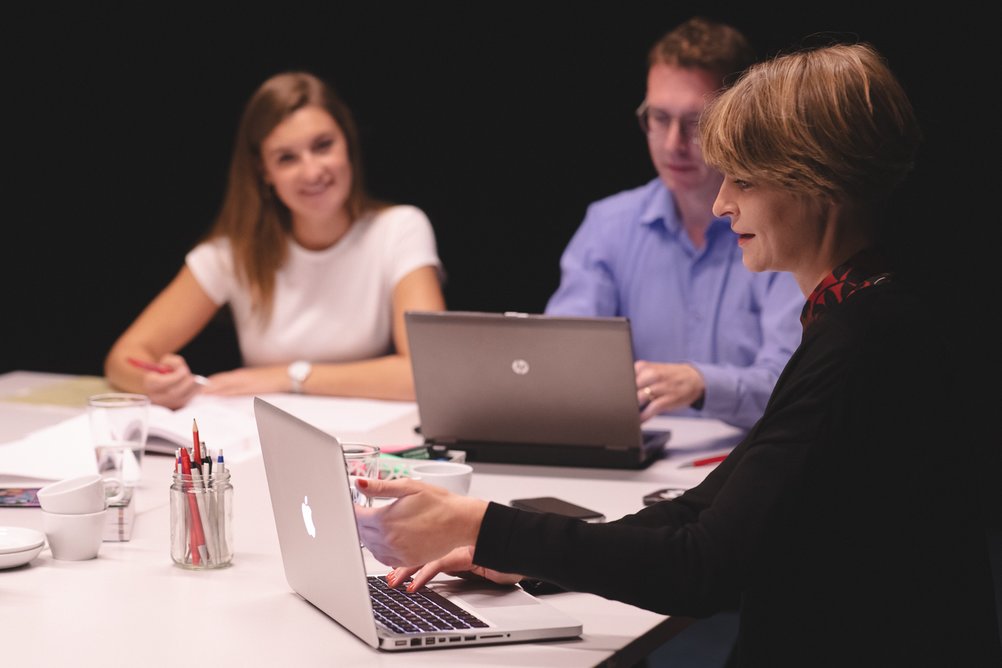 Drei Personen sitzen mit Laptops und Notizmaterial an einem Tisch und halten eine Sitzung ab.