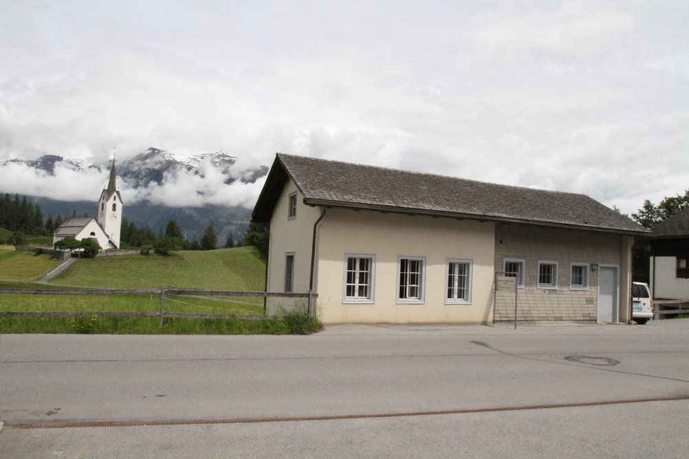 Das unscheinbare Gebäude im Vordergrund ist die historische Telefonzentrale von Versam. Im Hintergrund sind eine Kirche und hohe Berge zu sehen.