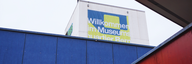 Architektur vor dem Museum für Kommunikation: verschiedenen Ebenen schneiden sich eine rote, eine blaue und eine dunkle Dachfläche. Dazwischen ein Ausschnitt Himmel und ein Liftturm mit einem Banner des Museumsquartiers Bern.  - vergrösserte Ansicht