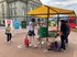 Ein Marktstand mit gelbem Dach aus Stoff. Interessierte Frauen mit grauen Haaren kommen zum Stand. Der Stand steht auf einem Platz in der Stadt.