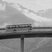 Schwarz-weiss-Fotografie eines Postautos. Das historische Bild zeigt ein Postauto, das auf einer luftigen Brücke über der Bergwelt zu schweben scheint.