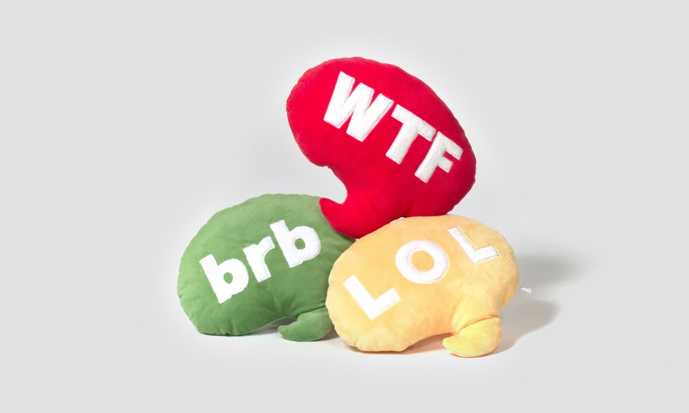 Three emoji pillows in the form of speech bubbles in different colours lie on a pile. On the pillows are the texts WTF, brb and LOL.