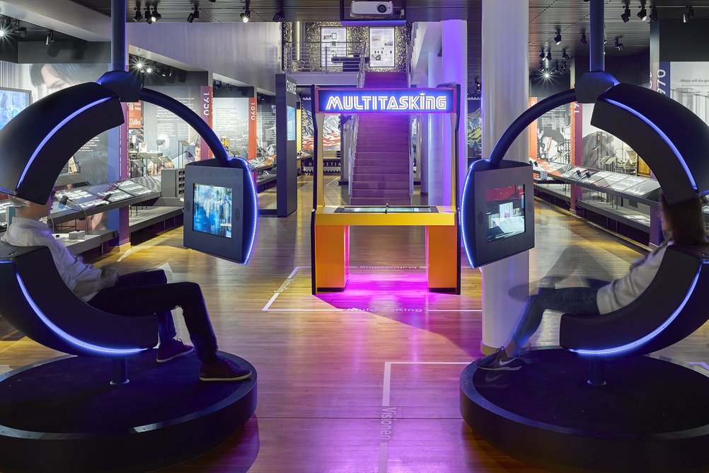 Ein Blick in die Zone Change in der Kernausstellung des Museums für Kommunikation. Im Vordergrund sind zwei futuristische Drehstühle zu sehen, im Hintergrund eine Spielstation.