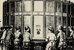 Frauen und Männer in Kleidung des 19. Jahrhunderts mit Kleidern, Anzügen und Hüten sitzen um eine Art zirka drei Meter hohe Tonne und schauen durch kleine Löcher in die Tonne hinein. - vergrösserte Ansicht