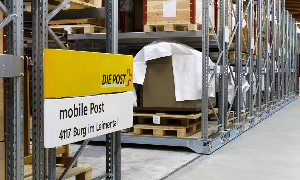 Ein Hochregallager in einem Museumsdepot. In Paletten verpackt sind Sammlungsobjekte zu sehen, im Vordergrund ist ein Postschild zu sehen.