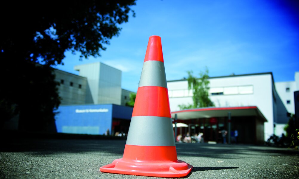 Auf asphaltiertem Boden steht ein orange-weisser Verkehrsleitkegel. Im Hintergrund sind die Gebäude des Museums für Kommunikation zu sehen.