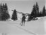Eine verschneite Landschaft mit Bäumen im Hintergrund. Vorne eine Spur von Skis im Schnee, eine Person steht in der Bildmitte mit Holzskis und einem dicken Stock in beiden Händen.