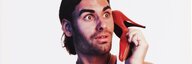 Ein junger Mann im Tanktop hält sich mit erstauntem Blick einen Absatzschuh ans Ohr als würde er telefonieren. Es ist ein Swisscom-Plakat von 1998. - vergrösserte Ansicht