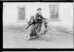 Schwarz-Weiss-Fotografie. Auf einem hölzernen dreirädrigem Fahrrad zwei Personen – ein Mann und eine Frau. Im Hintergrund eine Hauswand mit zwei vergitterten Fenstern.