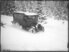 Die schwarz/weisse Fotografie zeigt eine winterliche Landschaft in waldigem Gebiet. Von links nach rechts führt eine Strasse, auf der ein Postauto mit Raupenantrieb über eine schneebedeckte Strasse fährt. Die aufgewirbelte Schneewolke zeigt, dass das Postauto verhältnismässig schnell fährt.