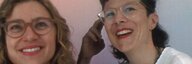 Zwei weiblich gelesene Personen lachen in Kamera vor neutralem Hintergrund. Beide haben gewelltes Haar bis zu den Schultern und beide tragen Brille. Die Person rechts fasst sich an den Kopf. - vergrösserte Ansicht