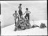 Aus dem Schnee ragt eine Steinpyramide in den Bergen, darauf stehen drei Männer mit nacktem Oberkörper.  Die Rucksäcke der Wanderer liegen daneben im Schnee.