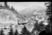 Schwarz-Weiss-Fotografie. Blick in eine Berglandschaft. Im Vordergrund des Bildes sind Bäume, dahinter ein kleiner Staudamm in dem Menschen baden. Im Hintergrund ein Hotel.