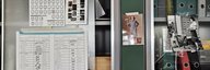 Eine Wand im Büro: In Schränken mit Glasscheiben stehen zahlreiche Ordner. An den Glasscheiben kleben Fotos und Blätter mit Tabellen. - vergrösserte Ansicht