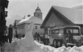 Auf der Schwarz-Weiss-Fotografie ist eine Situation in einem Dorfzentrum ersichtlich. Mehrere Häuser säumen die zentrale Strasse, die diagonal von rechts nach links führt. Es liegt viel Schnee. Im rechten Bild ist ein Postauto mit drei Achsen perspektivisch von hinten ersichtlich, das an der Rädern mit Schneeketten ausgerüstet ist. Vor dem Postauto steht ein mit einem Pferd bespannter Schlitten. Auf der Strasse befinden sich diverse Personen.