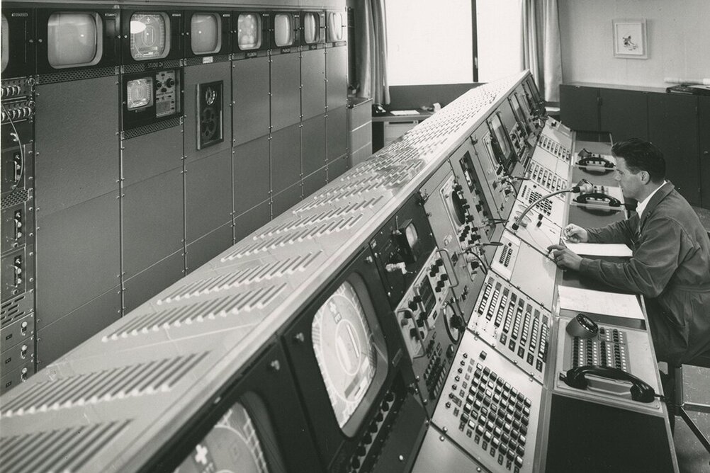 Une image historique d'un grand centre de contrôle avec des écrans et des panneaux de contrôle. Un employé est assis devant la console.