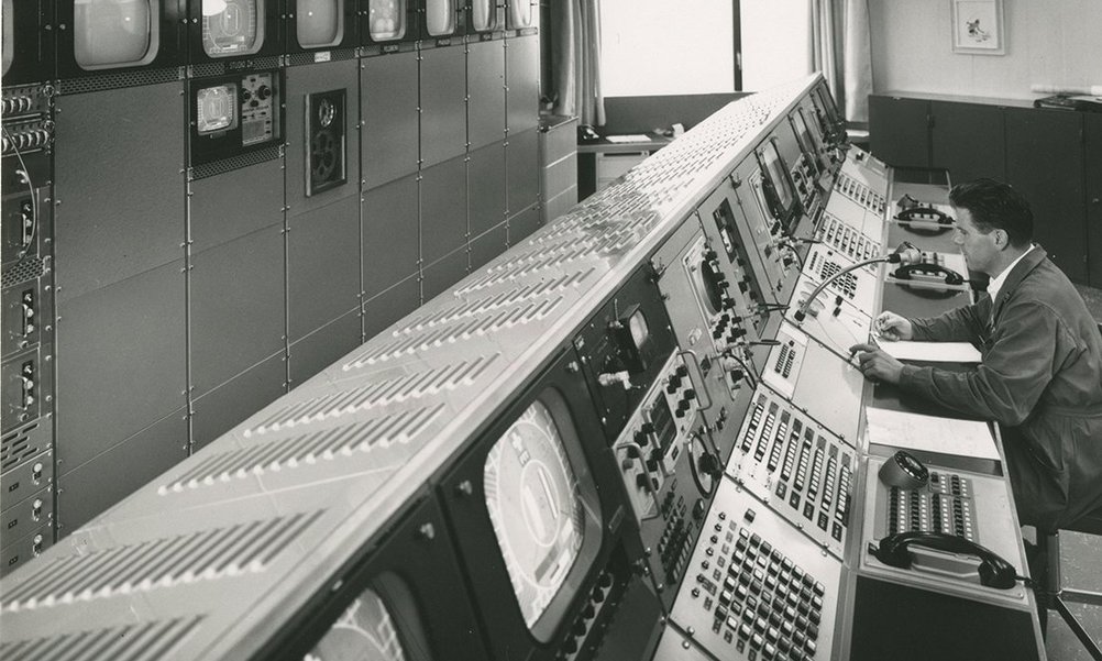 Ein historisches Bild einer grossen Schaltzentrale mit Bildschirmen und Schaltpulten. Ein Mitarbeiter sitzt vor dem Pult.