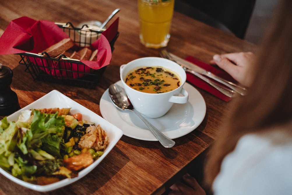 Über die Schulter einer Person blicken wir auf den Mittagstisch mit Salatteller und Suppe.