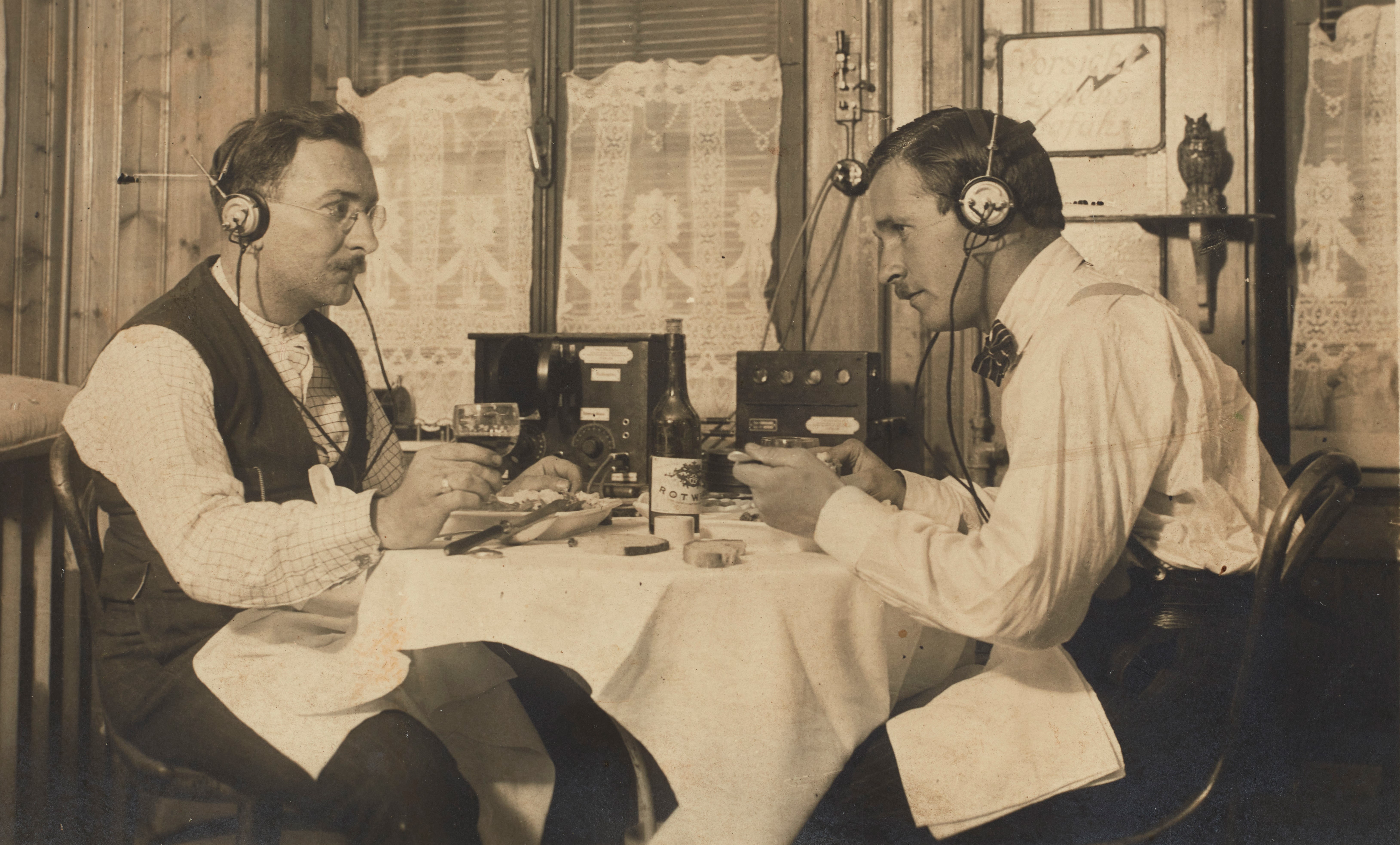 Die historische schwarz-weiss Aufnahme zeigt zwei gutgekleidete Herren an einem Tiisch mit weissem Tischtuch am Mittagessen. Auf dem Tisch steht ein Radiogerät, beide tragen Kopfhörer. - vergrösserte Ansicht